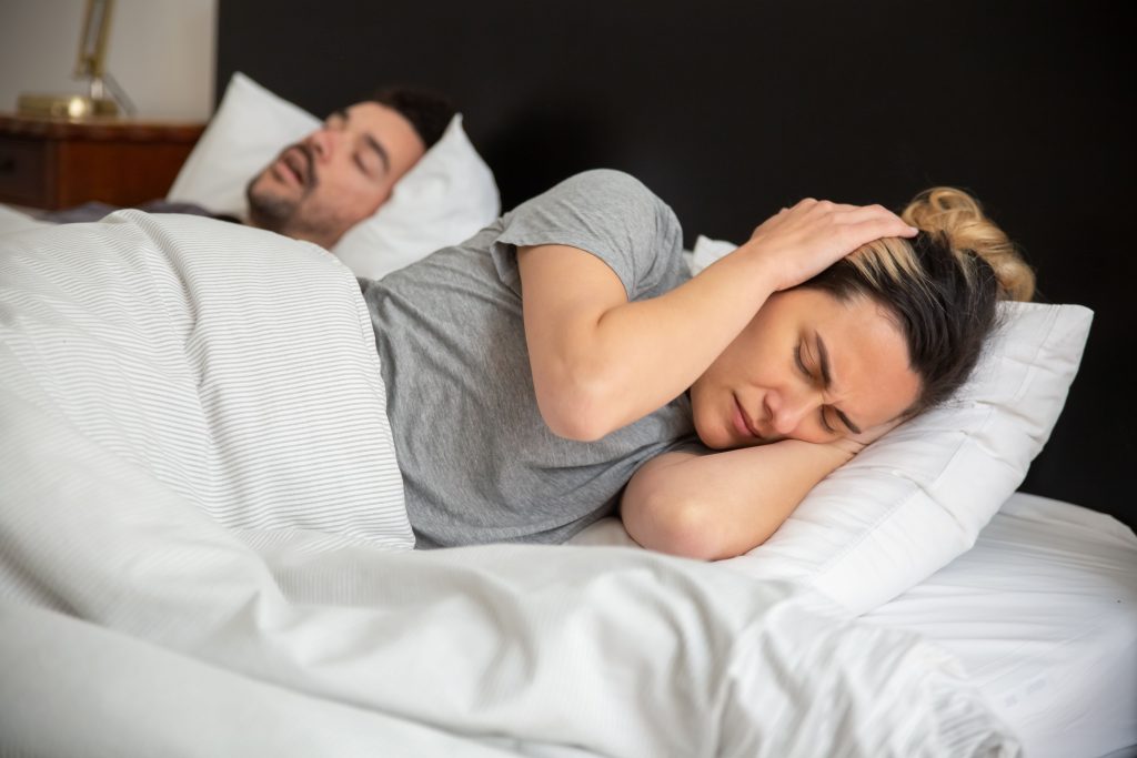 Is Snoring Bad? A woman looks frustrated in bed as her partner snores loudly, highlighting sleep disturbance issues.