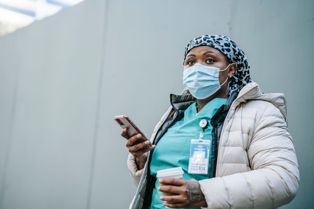 Nurse wearing mask and uniform using smartphone during outdoor coffee break.