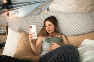 Mother uses smartphone while holding her newborn in bed, surrounded by pillows and cozy decor