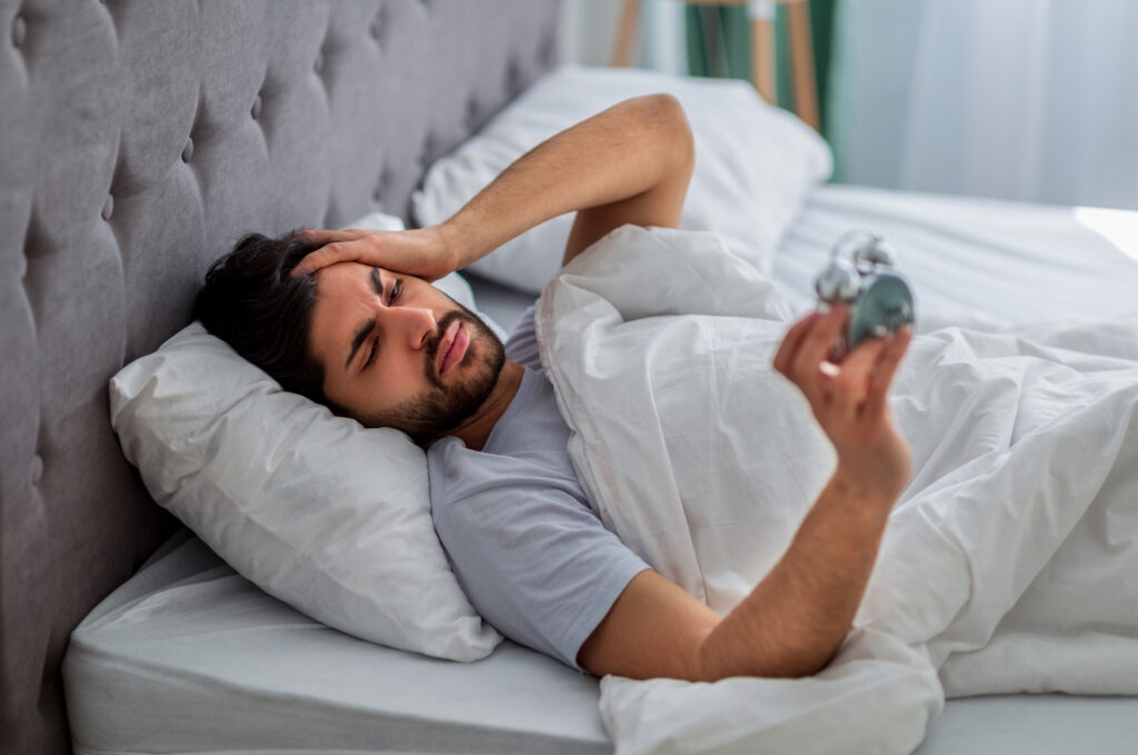 Man waking up unhappy and holding his head while looking at an alarm clock.