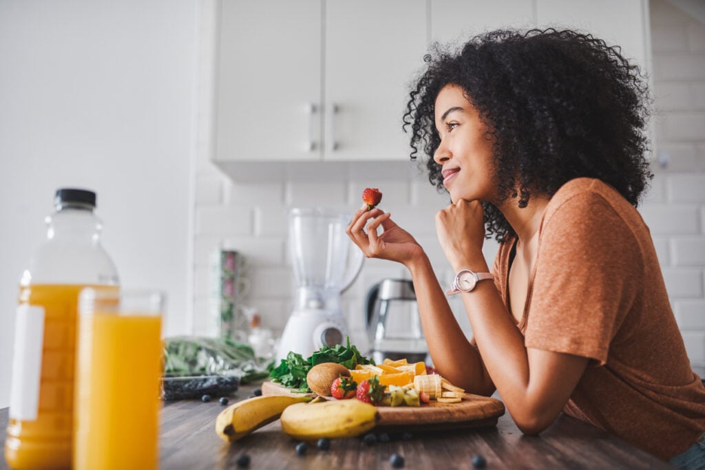 A young woman smiles while holding a strawberry in one hand with a plate of healthy food in front of her on the kitchen table