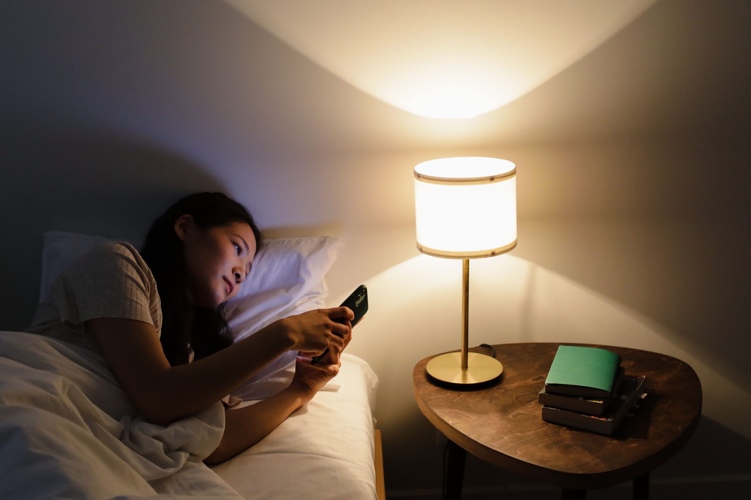 Light and Sleep A woman relaxes in bed, using her smartphone beside a lit table lamp in a cozy room