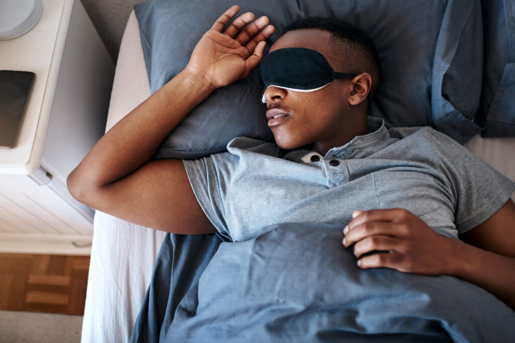 A young man wearing a night mask and sleeping in his bed at home