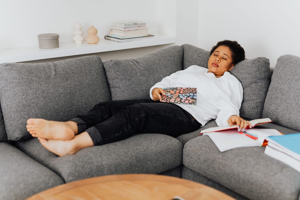 A young woman resting on a cozy grey sofa with open books around her, appearing tired after studying