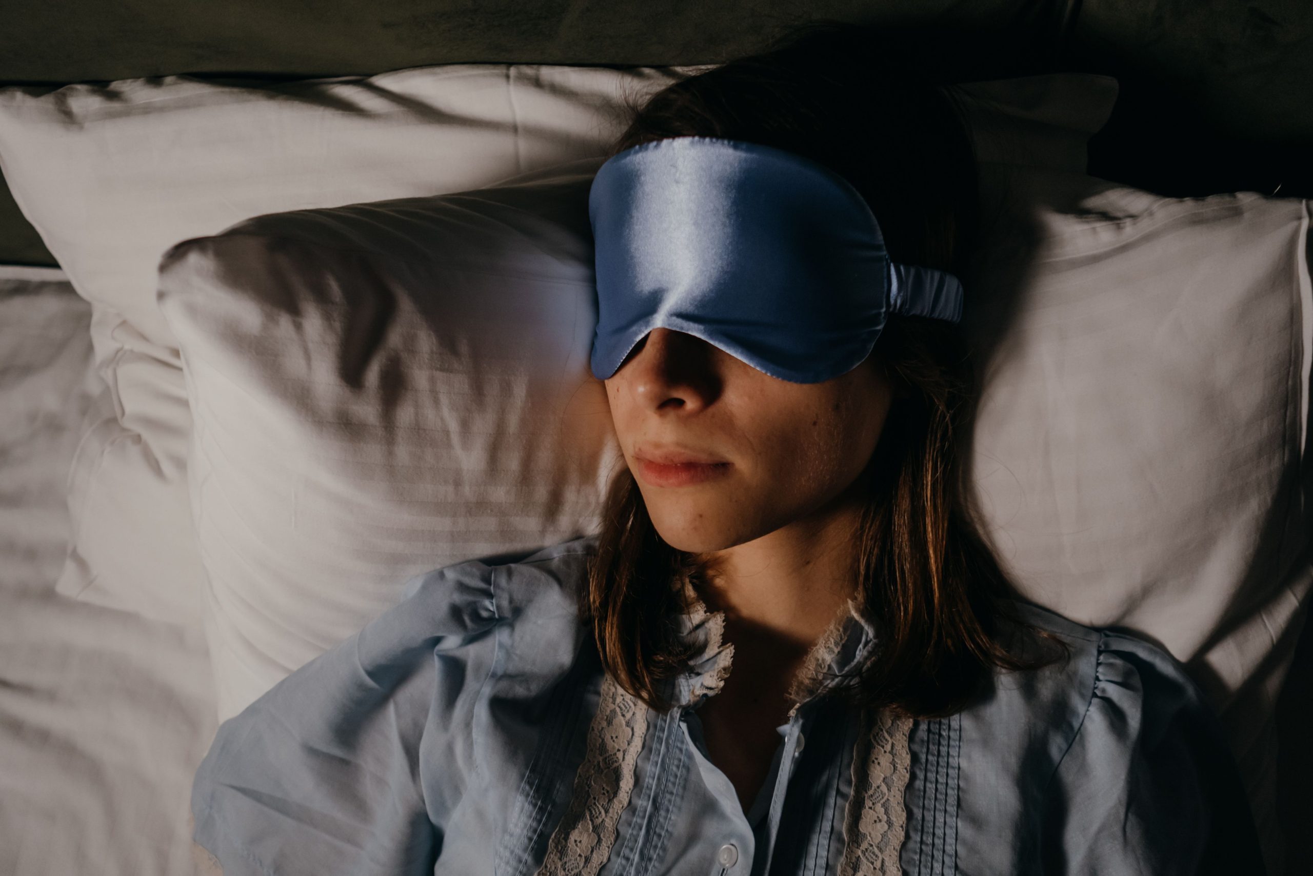 How Much Sleep Do Adults Need? Woman sleeping on bed wearing a soft blue eye mask for peaceful rest.