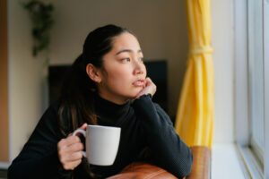 A young woman is sitting on a sofa staring out of the window and enjoying a hot drink at home in the living room.