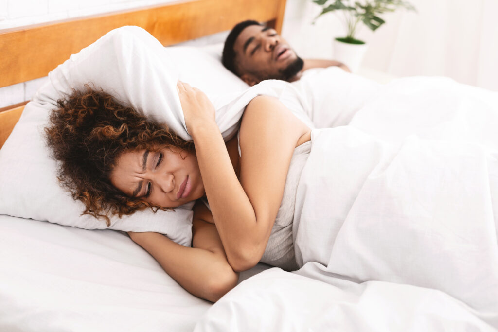 A woman covers her ears as her partner snores loudly in the bed next to her.