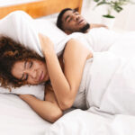 A woman covers her ears as her partner snores loudly in the bed next to her.