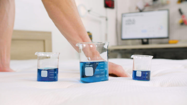 Sleep Foundation tester pressing the Silk & Snow Organic Mattress beside beakers of blue liquid during motion transfer testing