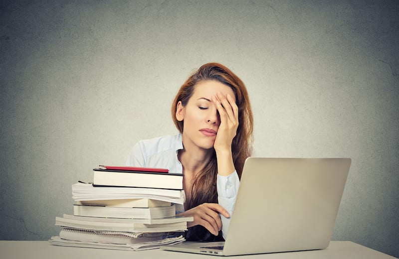 A tired woman sits at her desk with an open laptop and pile of books