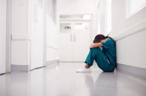 Stressed And Overworked Nurse Wearing Scrubs Sitting On Floor In Hospital Corridor