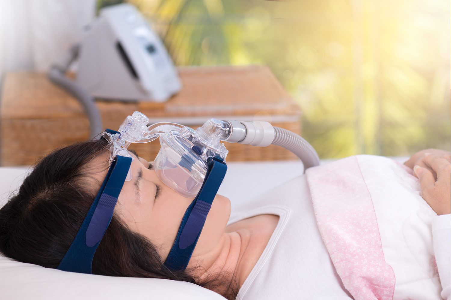 Happy and healthy senior woman wearing cpap mask sleeping smoothly without snoring on her back with blurred CPAP machine in background.Obstructive sleep apnea therapy, close up side view.
