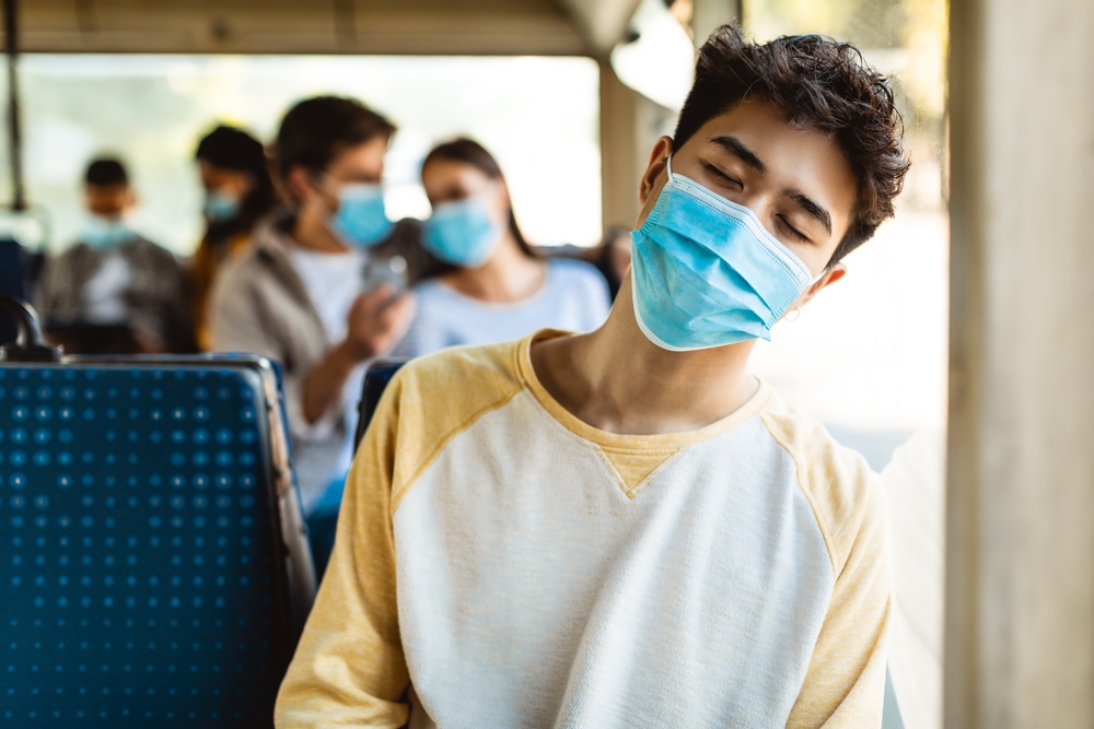 A young man wearing a face mask, rests his head on the window of a bus