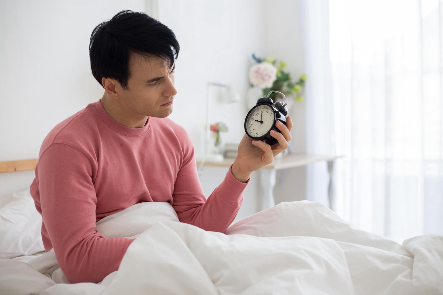 Man sitting on bed holding alarm clock. Sleep not wake up