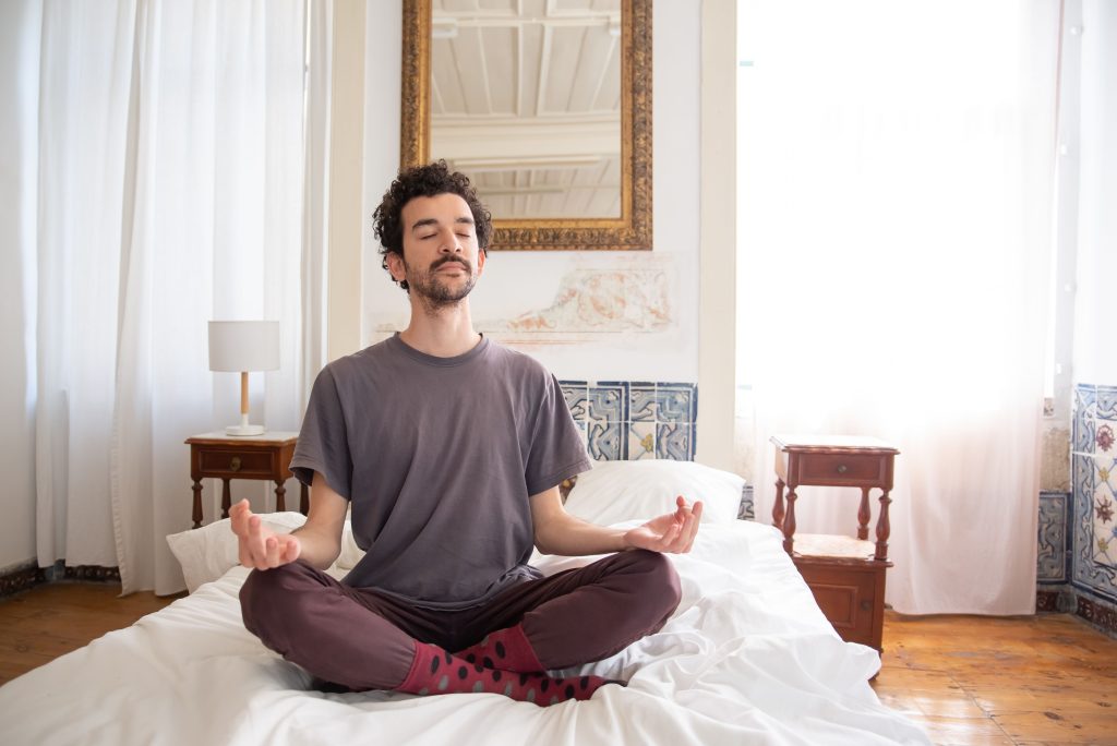 A man meditates calmly on a bed in a sunlit room, embracing tranquility and mindfulness.
