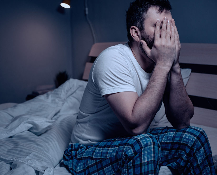 A distressed man sits on the edge of a bed with his face in his hands