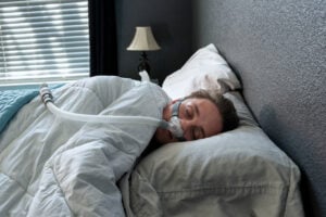 A man sleeps with a CPAP machine in bed. The machine helps him breathe better by delivering air pressure through a mask, improving sleep quality and reducing snoring