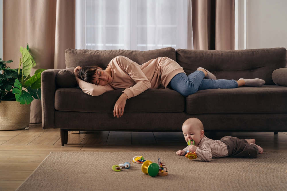 young mother suffering from post partum depression rests on a couch while her baby crawls on the floor